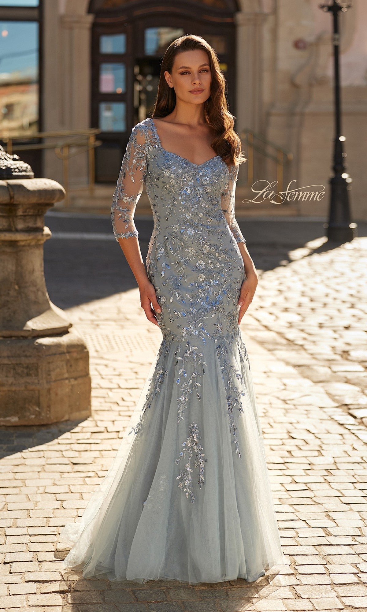 A woman stands outdoors on a cobblestone street in front of a historic building, wearing the La Femme 31799 long formal dress—light blue mermaid style with sheer sleeves and floral beaded embroidery.